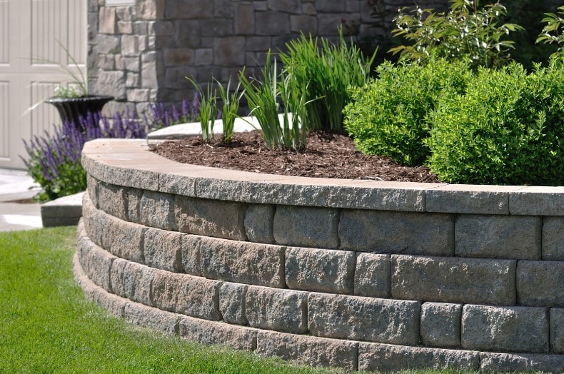 A retaining wall in a well maintained garden with green shrubbery planted on top, not failing as it was built by a professional.